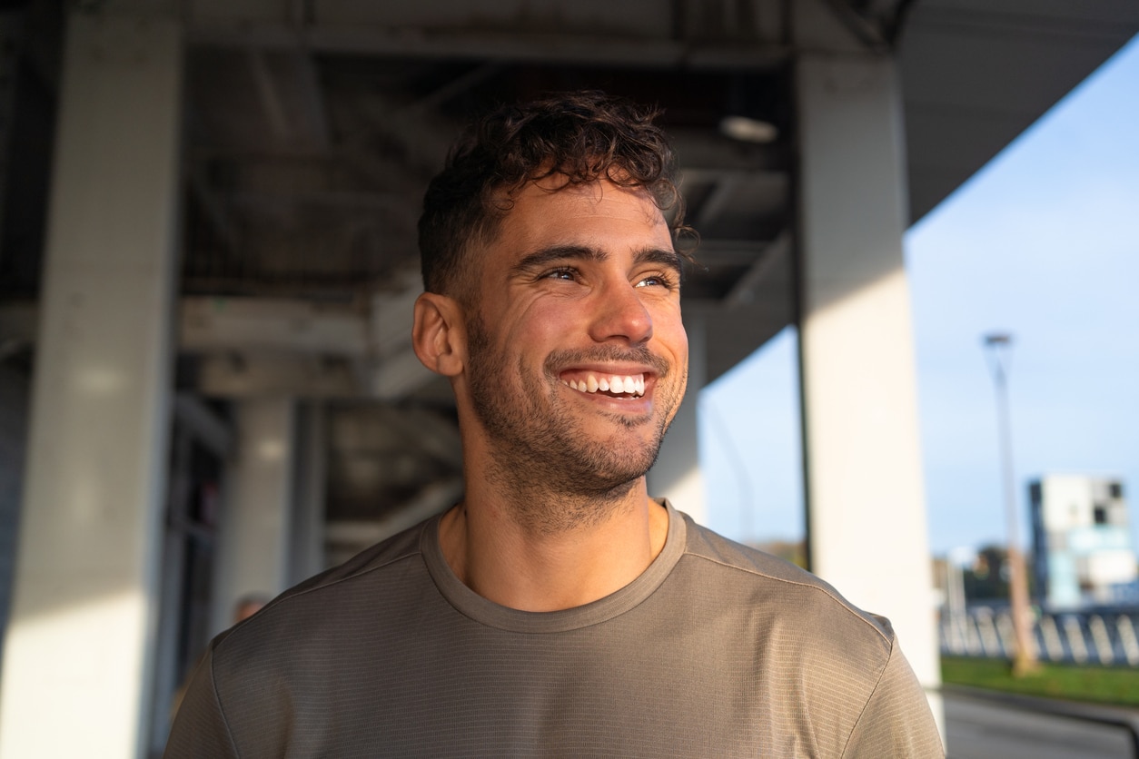 Smiling man outdoors under a sunny sky wearing a casual shirt in an urban setting.